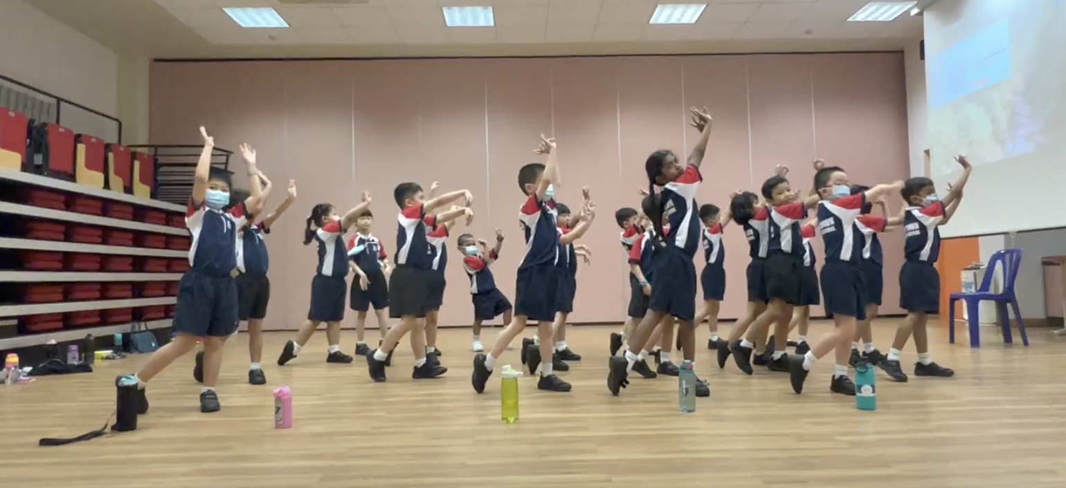 Young students in navy and red uniforms dance with arms raised in a school hall, wearing face masks.